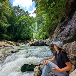Hiker overlooking the cascading white waters of Aranjuez River with a tall waterfall in the background, Boquerones Hike, Bajo Caliente, Puntarenas.