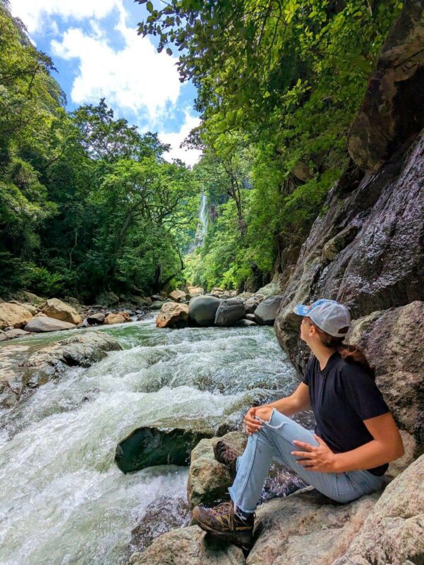 Hiker overlooking the cascading white waters of Aranjuez River with a tall waterfall in the background, Boquerones Hike, Bajo Caliente, Puntarenas.