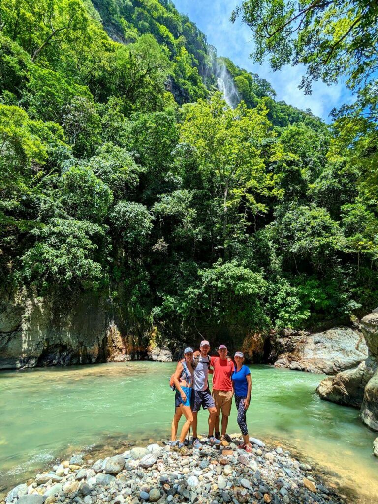 Group of hikers posing on riverside rocks during Boquerones Hike, Bajo Caliente, Puntarenas, with the Aranjuez River and mist from a seasonal waterfall in the backdrop.