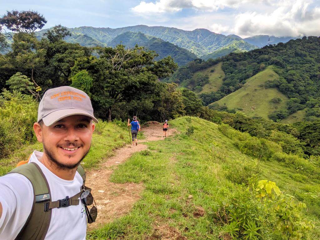 Hiker in Link Expeditions hat takes a selfie with fellow hikers and sunlit mountain tops in the background during the Boquerones Hike, Bajo Caliente, Puntarenas.