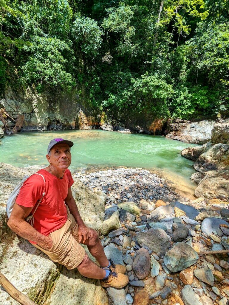 Hiker resting by the Aranjuez River at Boquerones, Bajo Caliente, with a deep natural pool and lush forest in the background.