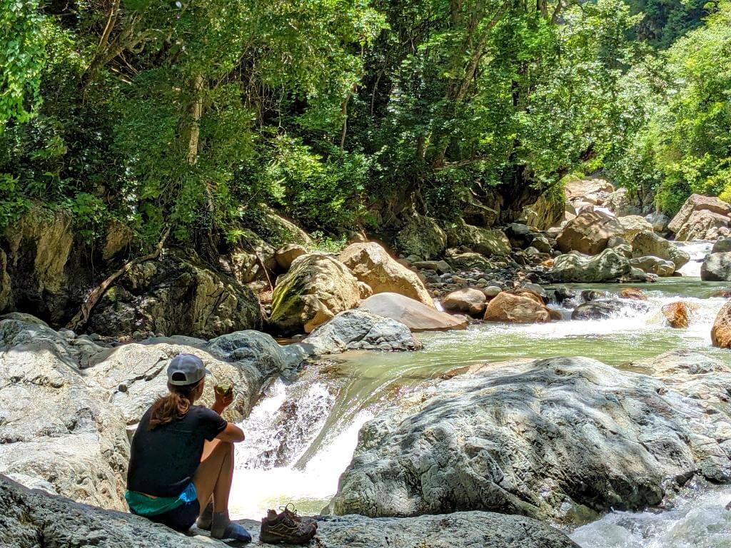 Traveler resting on a riverside rock while enjoying a snack, with the Aranjuez River flowing through a rocky canyon at Boquerones Hike, Bajo Caliente, Puntarenas.