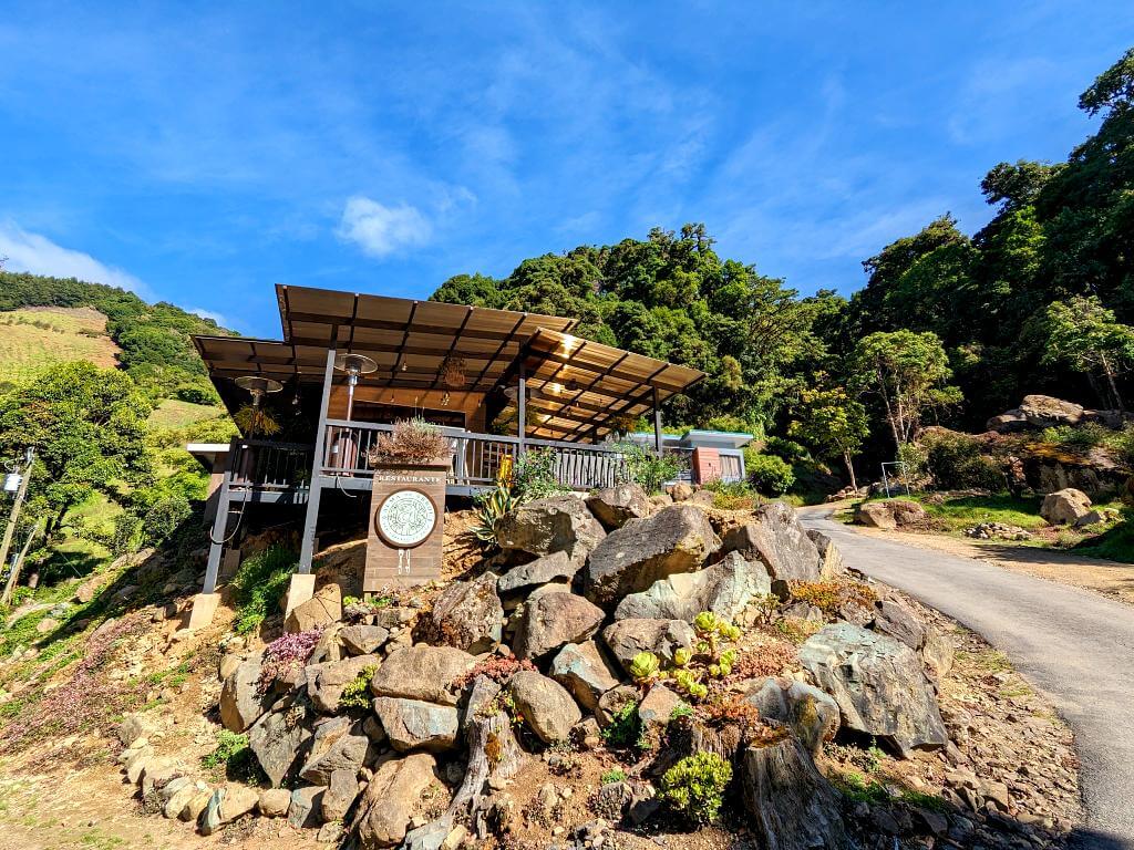 Entrance to Alma de Arbol restaurant in San Gerardo de Dota, with the restaurant visible in the middleground and the road leading to Batsu garden and forested hills in the background.