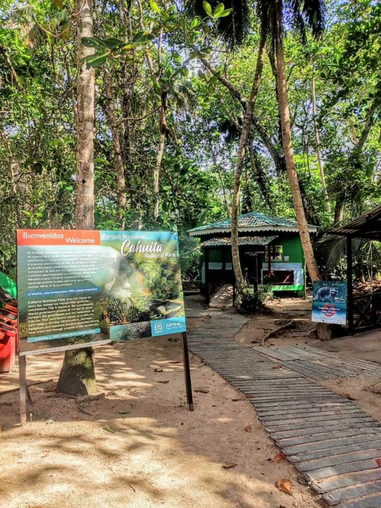 Entrance to Cahuita National Park featuring an informative sign, the ranger station, and lush greenery with numerous palm trees set on sandy soil.