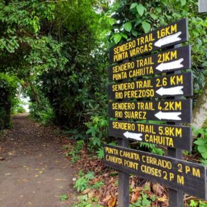 Signpost indicating trail distances at Cahuita National Park with a sandy flat trail surrounded by dense forest.