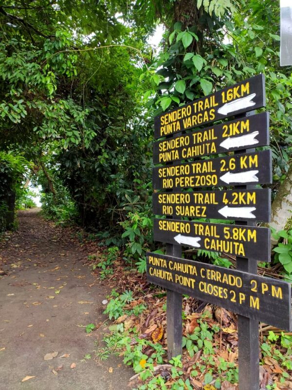 Signpost indicating trail distances at Cahuita National Park with a sandy flat trail surrounded by dense forest.