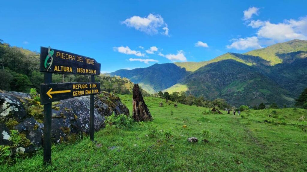 Signpost at 1850 meters above sea level reading "Piedra del Tigre," pointing towards Cerro Ena, located in a grassy area with impressive mountain views along the Cerro Ena Trail.