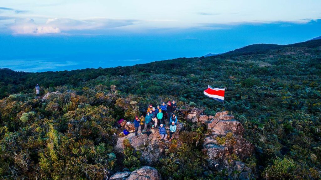 Aerial view of the mountain top of Cerro Ena captured by a drone, showcasing the vastness of the mountains, the Costa Rican flag fluttering proudly atop Cerro Ena, and a group of people marveling at the panoramic sunrise views.