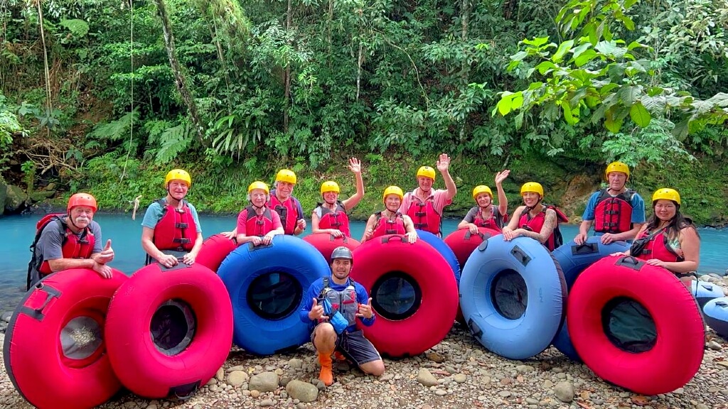 Large group of 11 adult travelers geared up for Rio Celeste tubing adventure, standing by the river's vibrant blue waters with their tour guide.