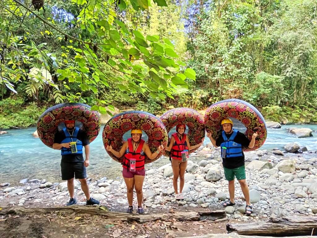 Four excited travelers holding inflatable tubes next to the turquoise Rio Celeste River, surrounded by lush green rainforest.