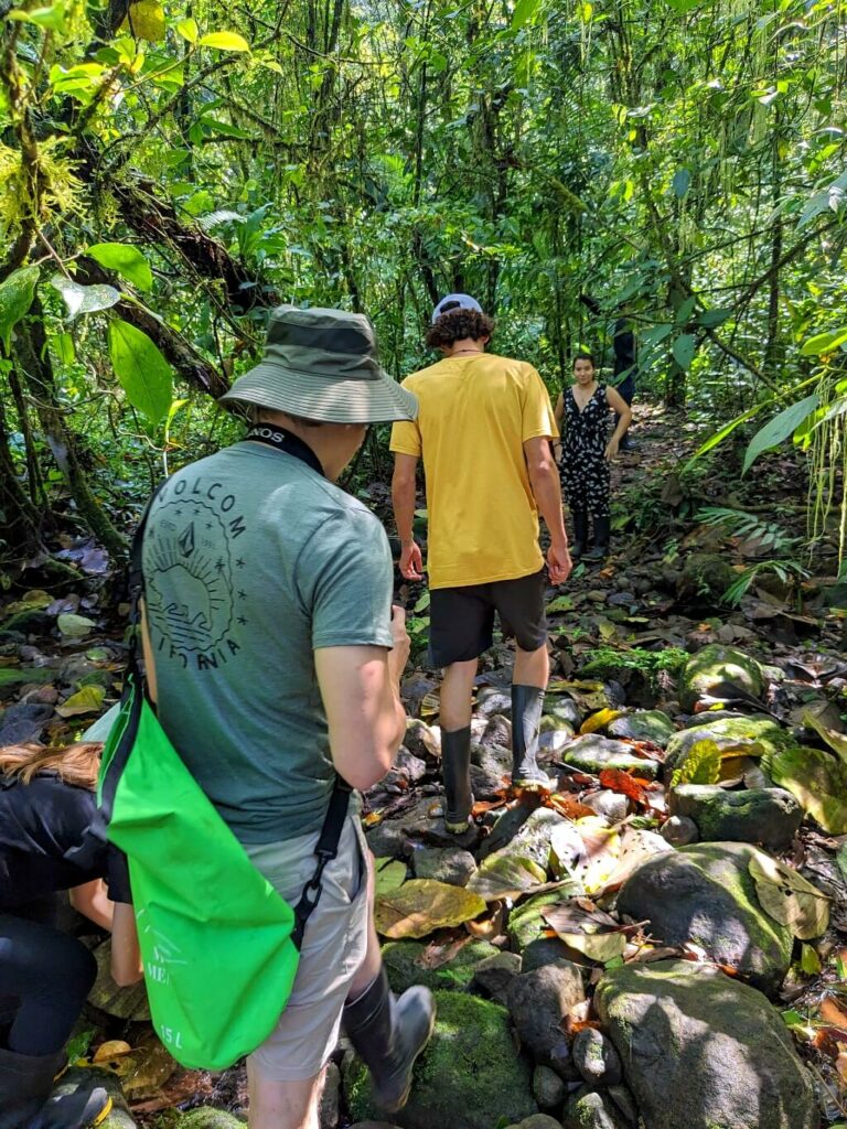 Two hikers in full gear (rubber boots, dry bags, cameras) navigate a rocky natural drainage on a trail in the Tapir Valley Reserve, Costa Rica.