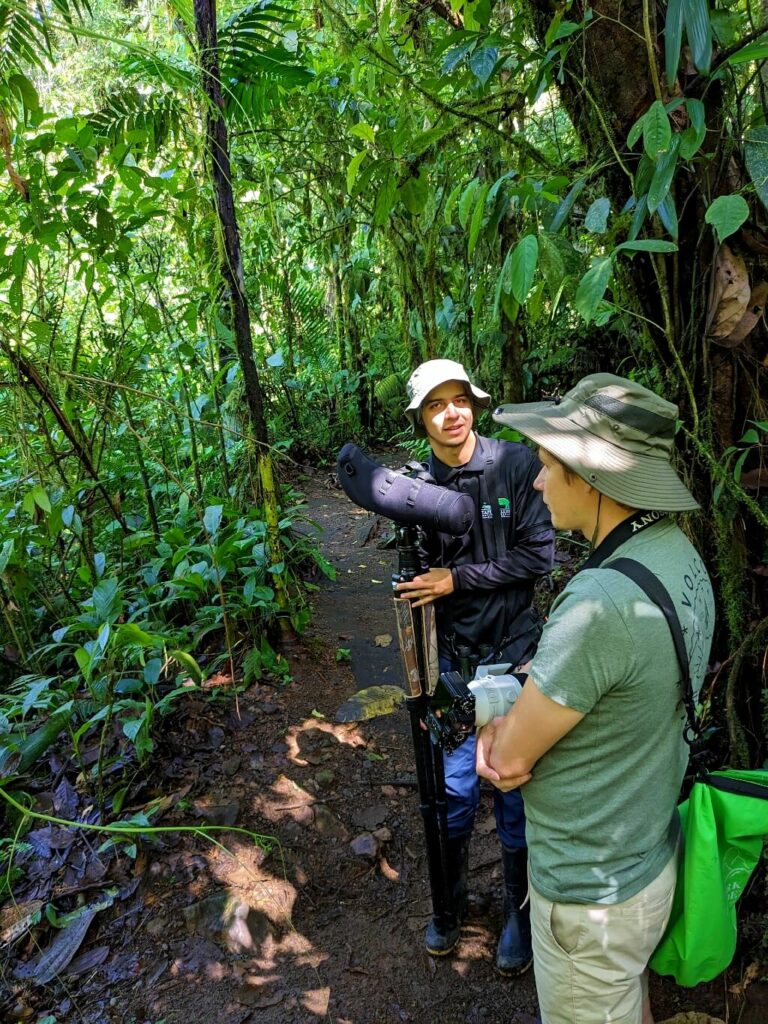 Tour guide in black long sleeve shirt holds a telescope while giving information to a curious traveler on a dirt trail in the rainforest.