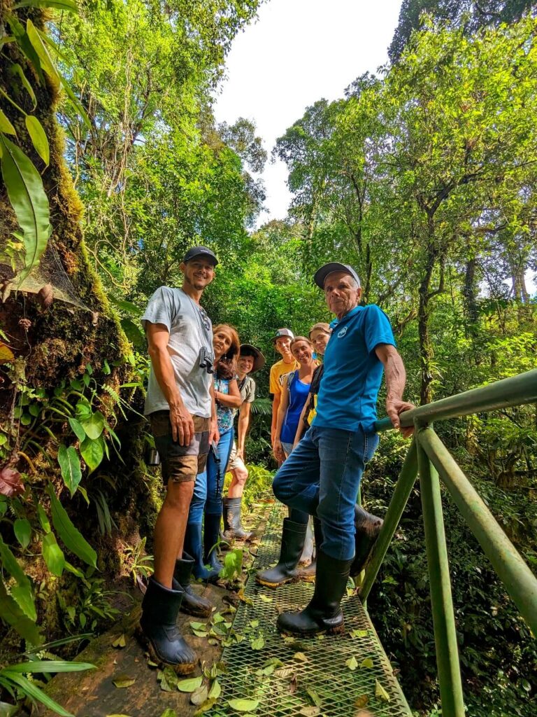 A group of Costa Ricans in protective rubber boots pose on a hanging bridge in the lush Bijagua rainforest, near Tenorio Volcano National Park and Rio Celeste. Cloudy skies and dense foliage surround them.