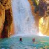Two people swimming and enjoying the falling water of the waterfall, with only their heads visible.