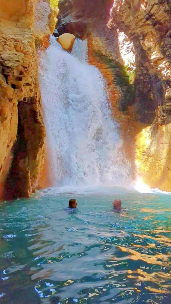Two people swimming and enjoying the falling water of the waterfall, with only their heads visible.
