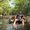 Relaxed man sitting on rocks in a river, surrounded by trees and nature.
