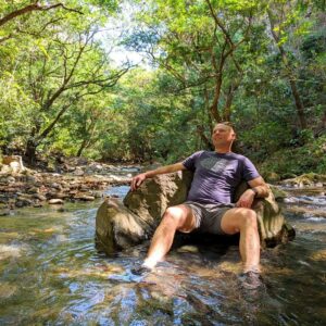 Relaxed man sitting on rocks in a river, surrounded by trees and nature.