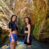 Two girls posing happily with the big rocks of the waterfalls and the blue water river in the background.