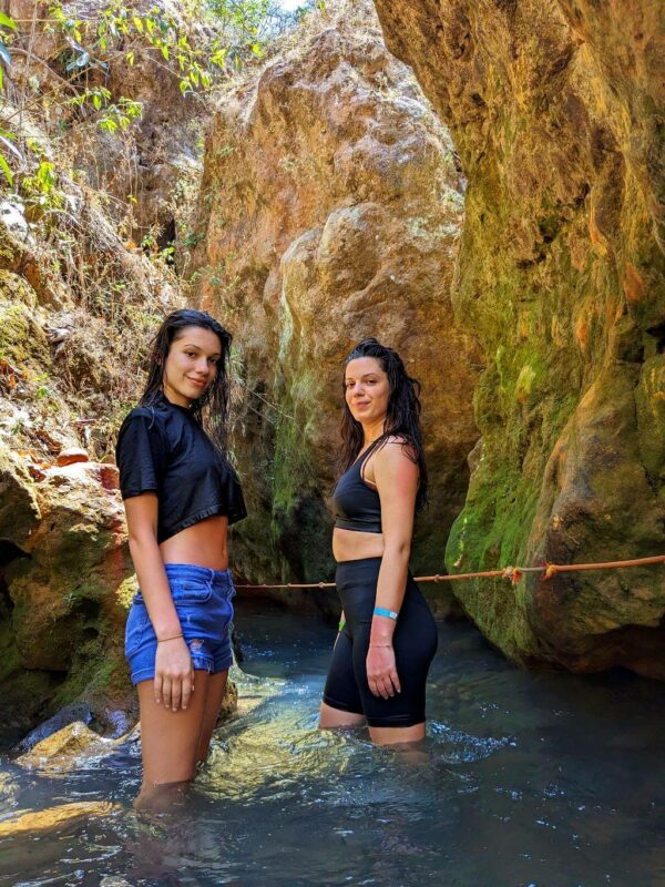 Two girls posing happily with the big rocks of the waterfalls and the blue water river in the background.