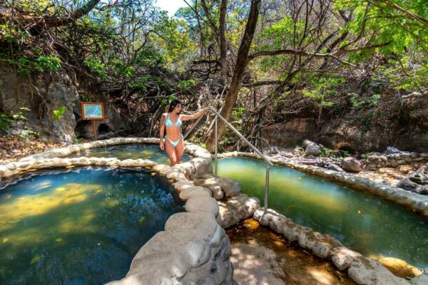 A woman enjoying hot springs in a natural landscape with crystal clear water and trees.