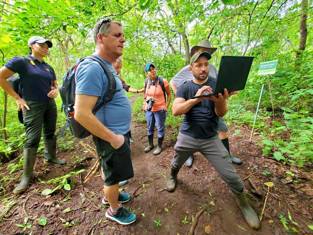 A group of people on a dry forest trail looks at a laptop with a biologist.
