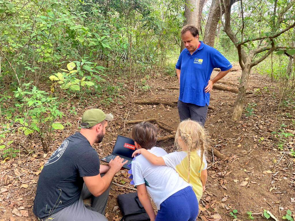 A biologist shows wildlife photos on a laptop to a father and two children on a trail.