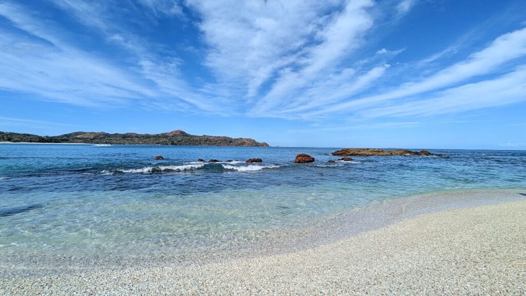 Clear blue Pacific Ocean and shell-covered white sand at Conchal Beach under sunny sky with soft clouds in Guanacaste, Costa Rica