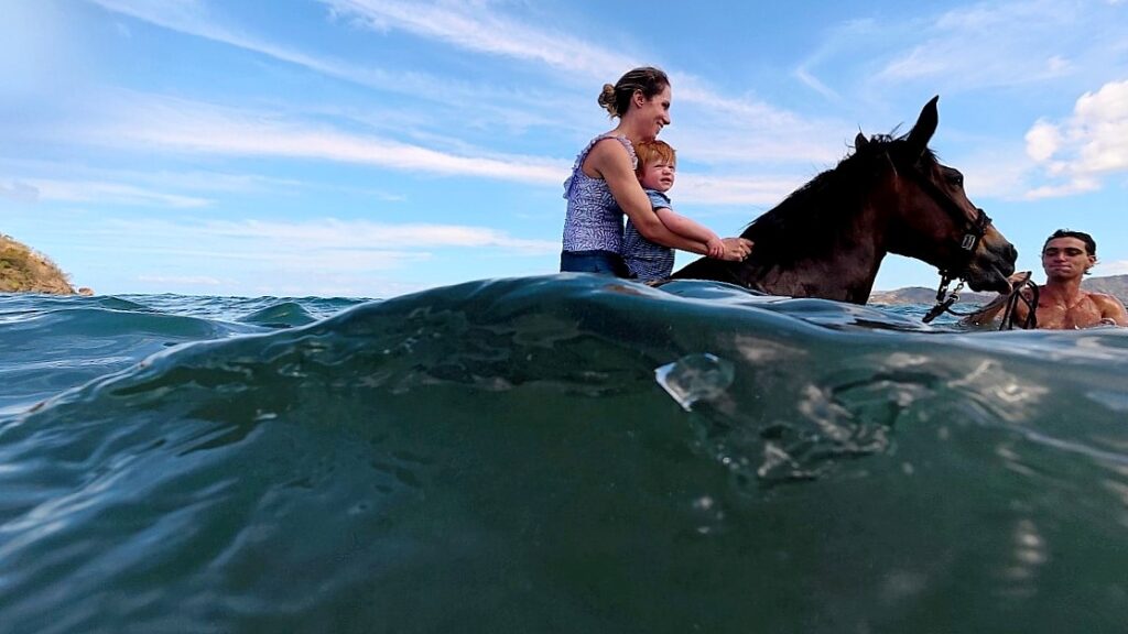 Mother and young child swimming on horseback with tour guide assisting in the ocean near Conchal Beach, Guanacaste, Costa Rica
