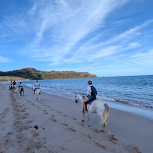 Group horseback riding on white sand toward Matapalo Beach with blue Pacific Ocean and sunny sky in Guanacaste, Costa Rica